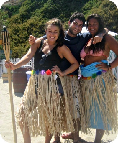 Three camp counselors standing together on the beach at Aloha Beach Camp's Hawaii summer camp program. The counselors are wearing grass skirts, colorful flower leis and holidng a Tiki Torch.