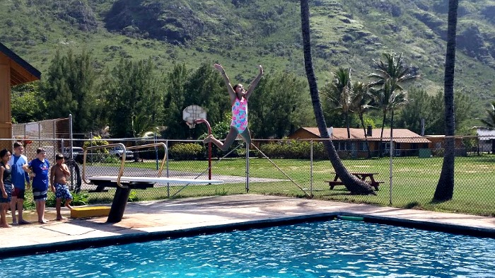 Female camper jumping off diving board in the clear blue swimming pool at summer camp in Hawaii