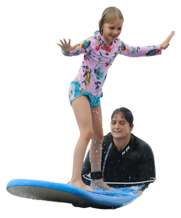 Camp counselor teaching a young female camper how to surf on her surfboard at Aloha Beach Camp Summer Camp in Hawaii.