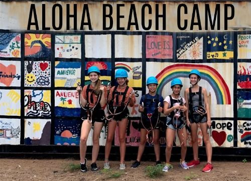 Five campers in ropes course gear (harnesses and helmets) standing in front of Aloha Beach Camp large tiled mosaic sign at summer camp in Hawaii.